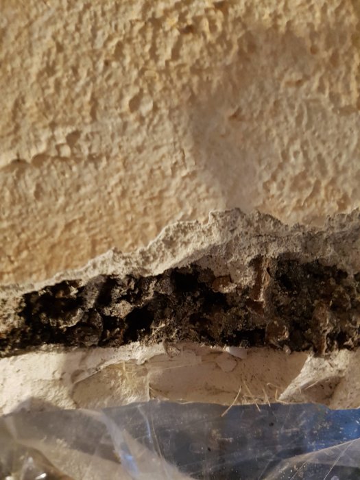 Skadad ventilation med lager av puts, svart material och vit bottenplatta, täckt av plast vid rivningsarbete.