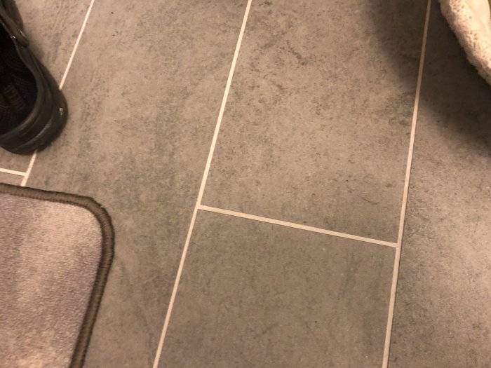 Grå klinkergolv med ljus fog som ser vit ut, del av en sko och mattkant syns.