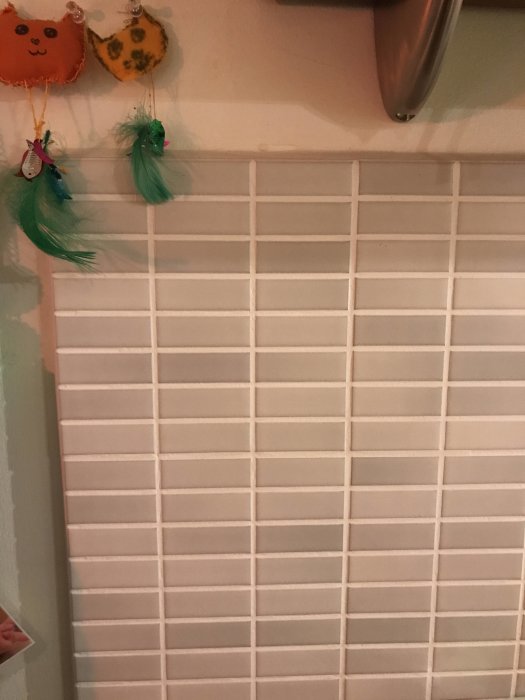 Vägg med vitt kakel och små fyrkantiga plattor, prydnadsföremål hängande ovanför.