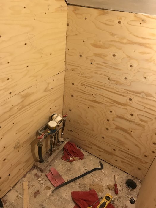 Hörn av ett rum under renovering med nya 15mm plywoodväggar och synlig rörgenomföring.