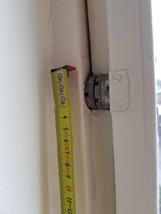 Måttband visar dimensioner på ett vitmålat pivåfönsters gångjärn i ett bostadshus.