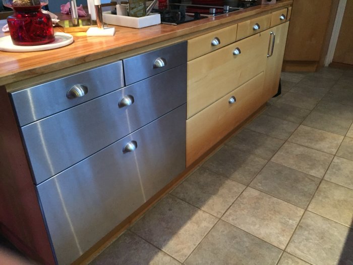 Köksskåp med en kombination av nya rostfria lådfronter och original träfärgade köksluckor under en bänkskiva.