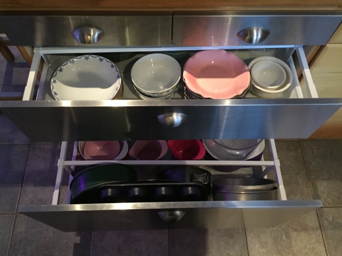 Kökslådor med rostfria fronter öppna, visande innehållet av skålar, fat och köksutrustning ordnat i skikt.