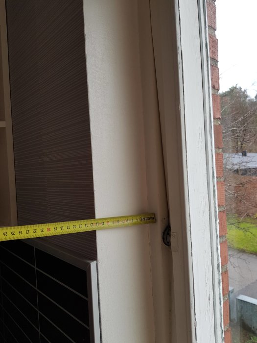 Måttband längs kanten på ett fönster som behöver renovering, med en synlig skruv och tegelvägg i bakgrunden.