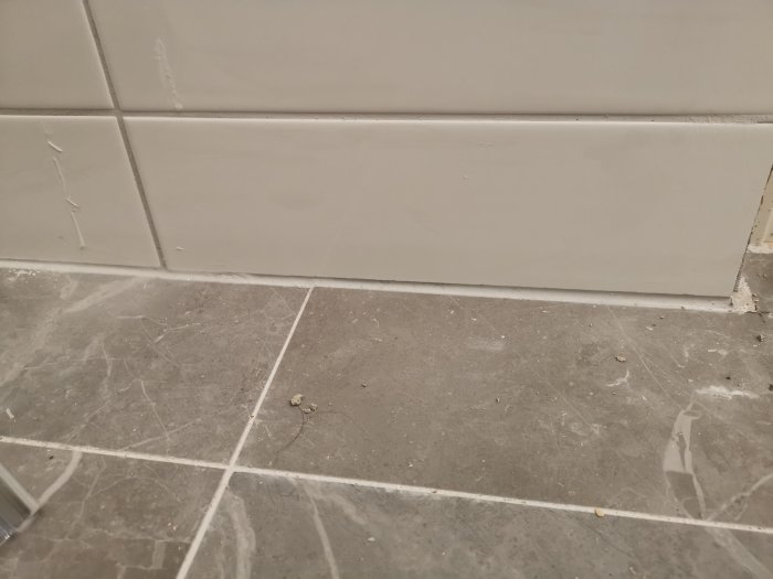 Gråa klinkergolv som möter vita kakelväggar med synliga ojämnheter och glipor vid övergången, nära hörnet av ett rum.