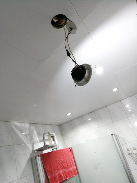 Öppen takinstallation med fläkt och spotlights nära en dusch i ett kaklat badrum.