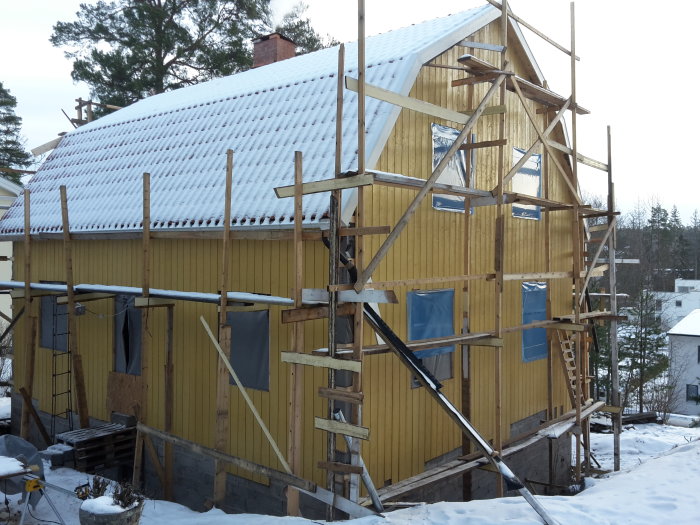 Hus under renovering med mansardtak, uppmurad skorsten och delvis täckt med vindpapp, omgivet av byggnadsställningar.