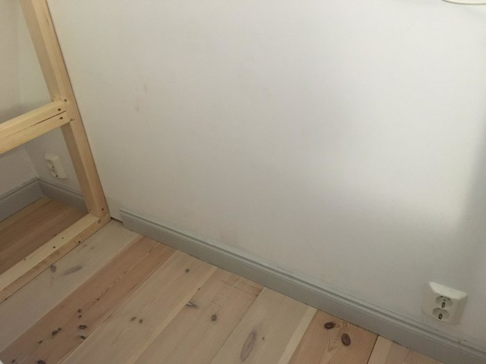 Hörn av ett rum med påbörjad sänggavelväggkonstruktion och ett vägguttag till höger.