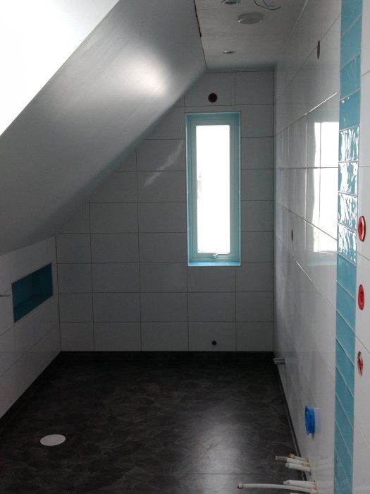 Renoverat badrum med vita kakelväggar, grått golv och lutande tak vid fönster.
