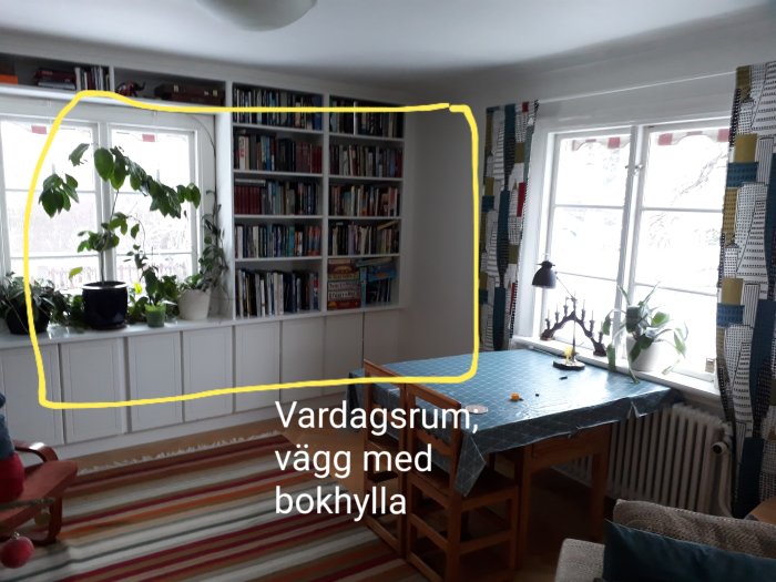 Vardagsrum med bokhylla längs väggen, markerad plats för ventilation och matbord framför fönster.