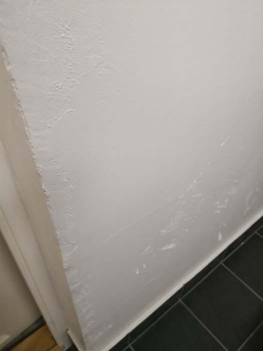 Ojämn och dåligt utförd putsning på en vit vägg ovanför svart klinkergolv.