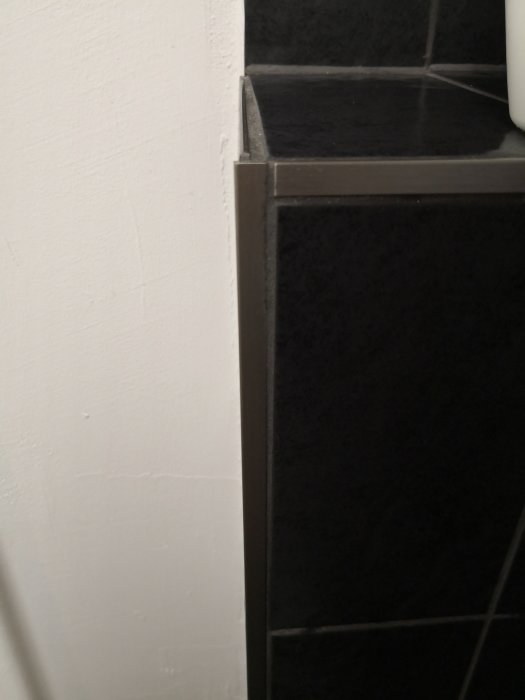 Hörndetalj i badrum med svarta kakelplattor, synliga fogar och en ojämn övergång till den vita väggen, vilket indikerar dåligt hantverk.