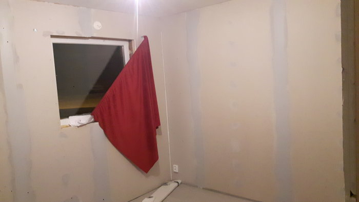 Sovrum under renovering med gipsskivor, spacklade skarvar och fönstersmyg samt ett rött tygstycke.