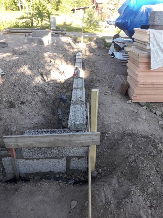 Grävd grund för verkstad med delvis murad stödmur och synligt dräneringsrör på byggplats.