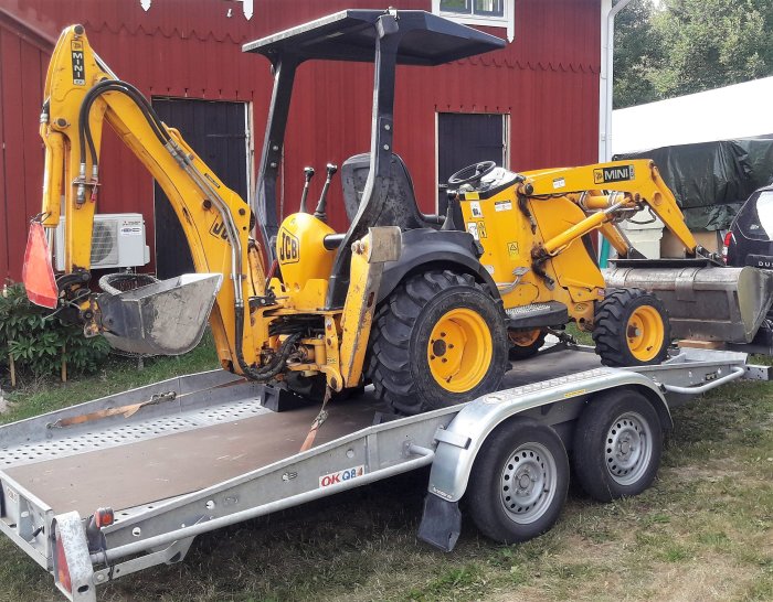 Liten gul JCB grävmaskin lastad på en släpvagn framför en röd byggnad.