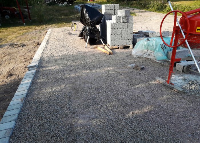 Grusplatta för byggprojekt med kantsten, betongblock, byggmaterial och en röd blandare på en byggarbetsplats.