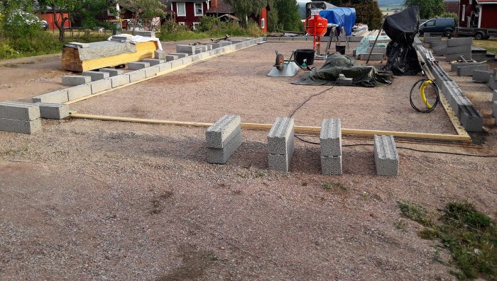 Grundläggning av en verkstad med betongblock och styrplankar på en grusbelagd yta, byggmaterial i bakgrunden.