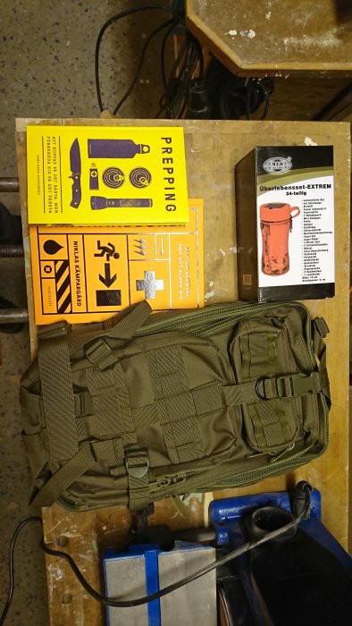 Preppingböcker och ryggsäck på verkstadsbord, samt en överlevnadsset-förpackning.