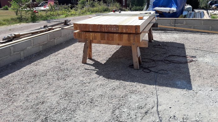 En arbetsbänk på en grusplatta vid en nybyggd stödmur med byggmaterial i bakgrunden.
