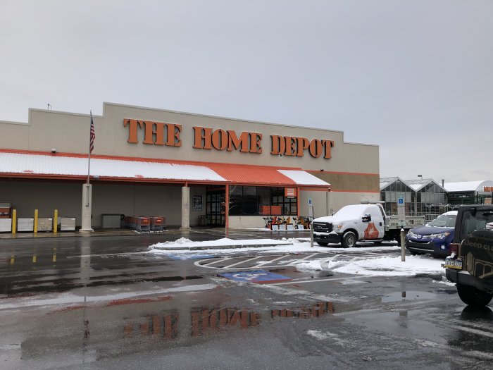 The Home Depot butik fasad med snö på marken och parkerade bilar under molnig himmel.