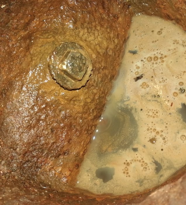 Mässingsplugg i hård yta med vattenansamling bredvid, svårtillgängligt för borrning.