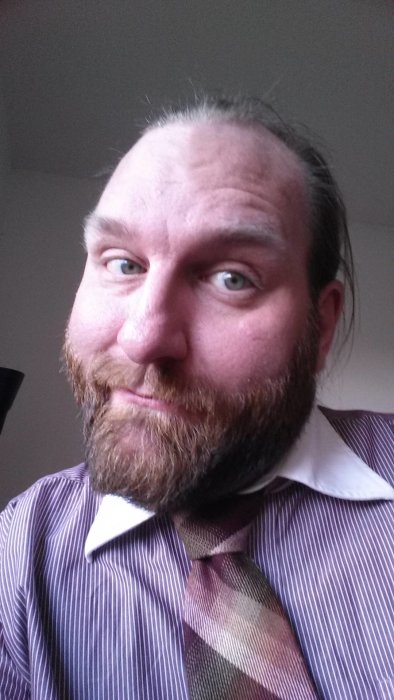 Man med långt hår och 25 mm skägg i randig skjorta och slips som ler.