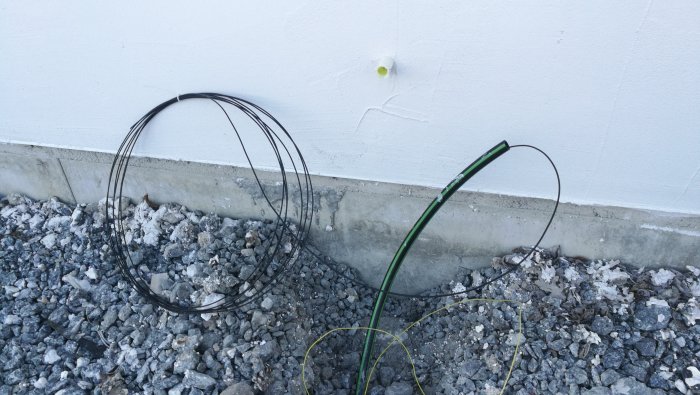 Framblåsta fiberkablar mot en vit husvägg med synliga rör och grus på marken.