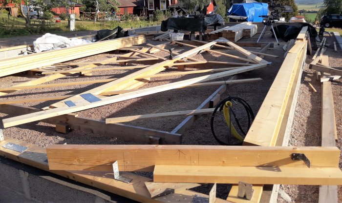 Byggplats med träsyll och takstolar under konstruktion samt byggmaterial och verktyg i förgrunden.