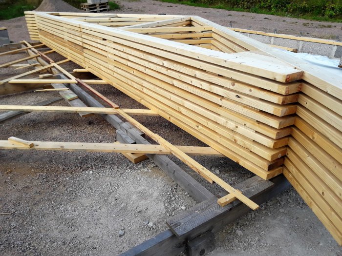 Stapel av timmer och takstolar förberedda för byggprojekt, upplagda på EU-pall och presenning.