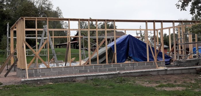 Pågående arbete på trästomme för byggnad med takstolmallar och byggmaterial skyddat med presenning.
