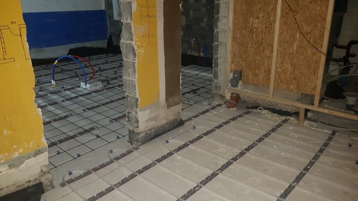 Nedervåning under renovering med isolering och nylagda rör för golvvärme före betonggjutning.