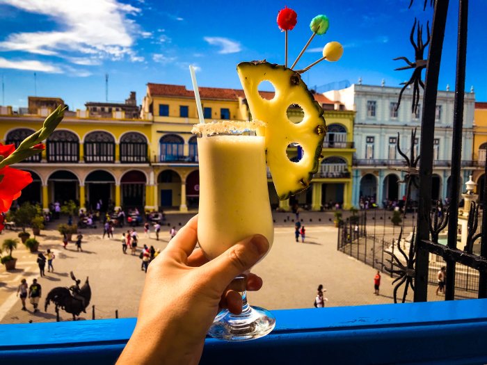 Hand håller en pina colada mot en färgglad karibisk torgbakgrund med människor och en häst.