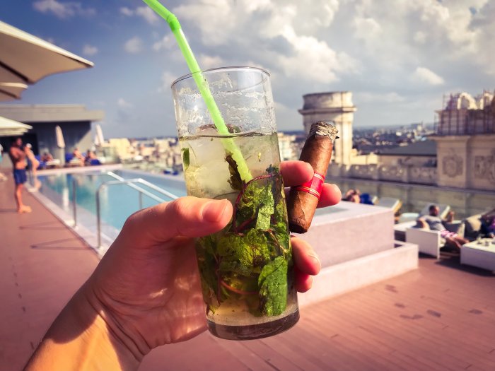 Hand som håller en mojito och en cigarr framför en pool på takterrass med människor och stadssilhuett i bakgrunden.