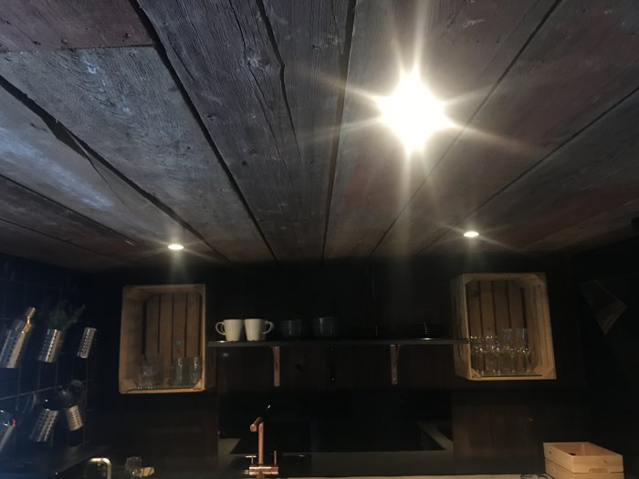 Mörkt kök med träpanel i taket och spotlights, svarta skåp, öppna hyllor och kopparfärgade detaljer.