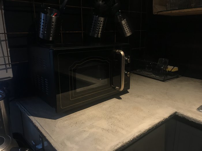 Svart mikrovågsugn på en köksbänk med kakelvägg och köksredskap i förvaringskärl ovanför.