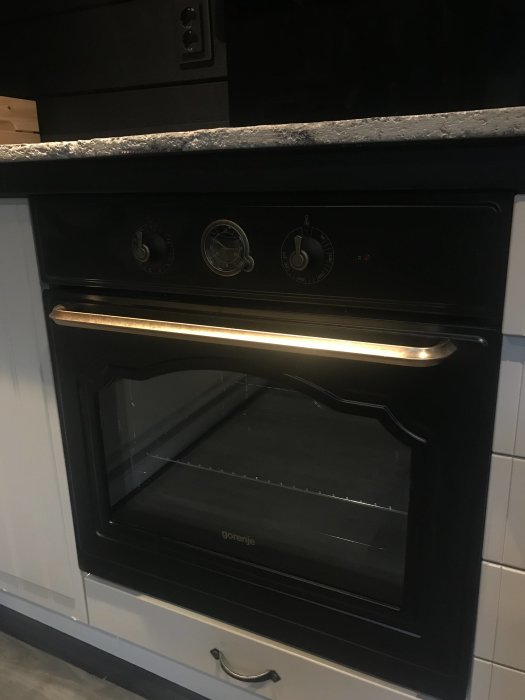 Svart inbyggd ugn i kök med rostfritt handtag och kontroller, omgiven av svarta skåp och stenbänkskiva.