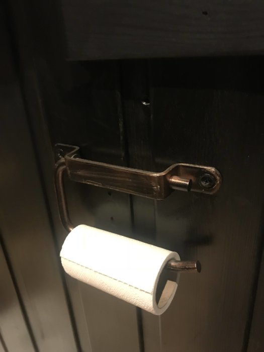 Toalettpappershållare av metall på en mörk trädörr, papperet är rättvänd.
