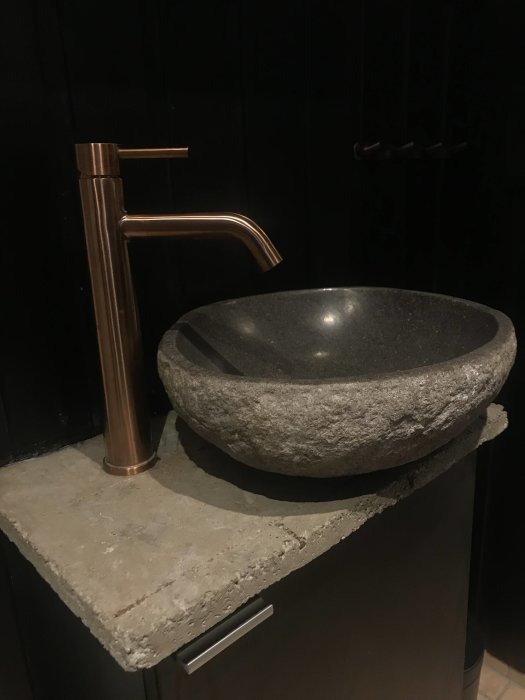 Modern köksblandare i borstad mässing och handfat i sten på en grov betongbänk mot en svart vägg.
