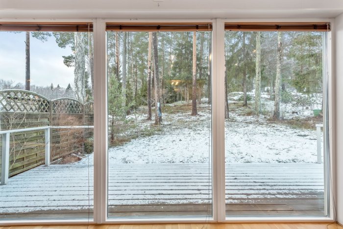 Utsikt från ett fönster över en snöbeklädd trädäck som gränsar till en sluttande skogstomt med snö och barrträd.