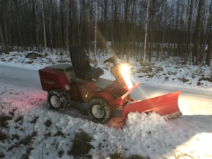 Röd traktor med plogblad framför plogar snö på en väg vid skymning.