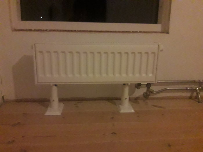Konvektor radiator element av vit färg monterat vid ett fönster, på ett trägolv i ett rum.