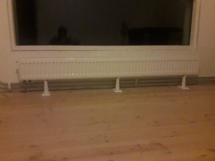Lång vit konvektorradiator monterad under fönster i ett rum med trägolv.