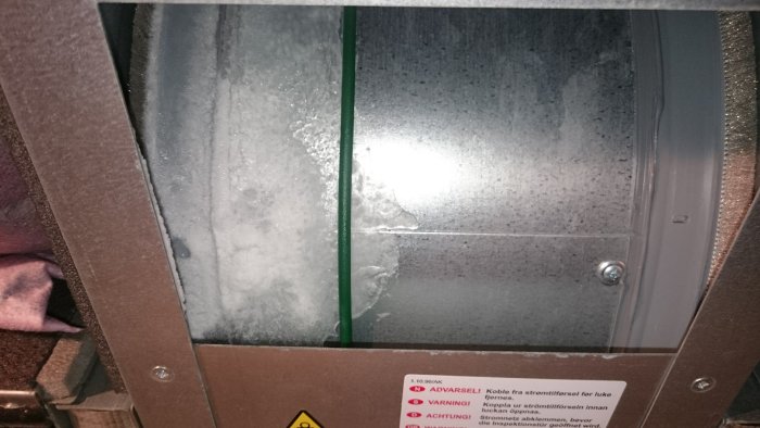 Isbildning på roterande trumma i ventilationssystem med synlig ishinna och varningsetiketter.