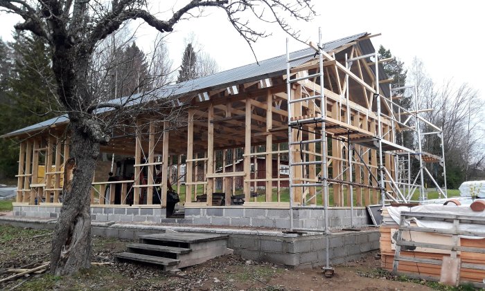 Byggnadsställning runt en påbörjad träkonstruktion av ett hus med material spridda runtom.