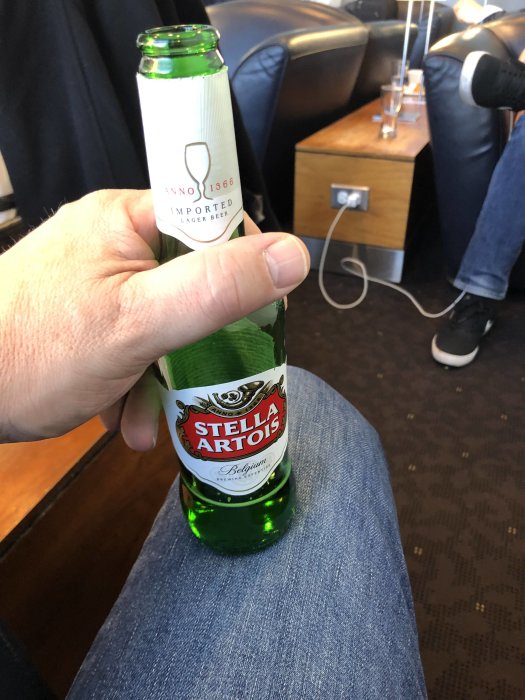 Hand håller en flaska belgiskt Stella Artois öl i en loungemiljö med flygplatskänsla.