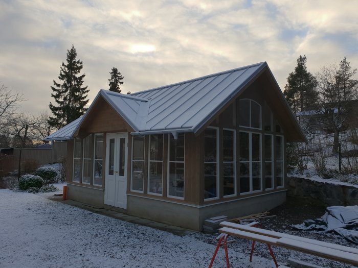 Snötäckt klickfalsat aluzinktak på ett hus med stora fönster och byggmaterial i förgrunden.