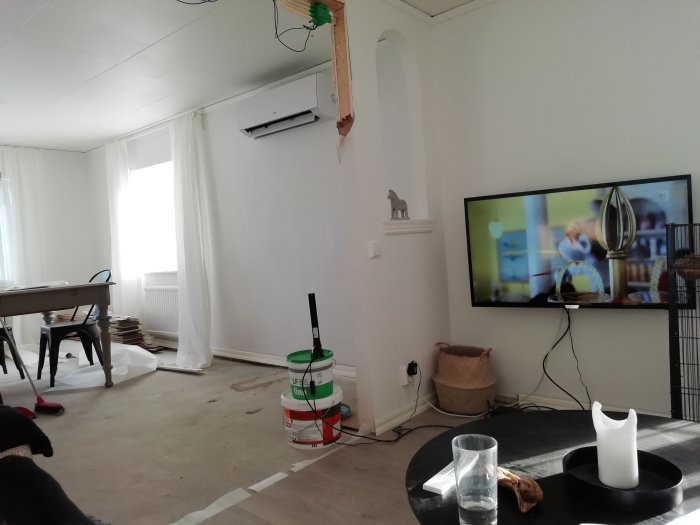 Vardagsrum under renovering med luftvärmepump, TV och diverse byggmaterial synliga.