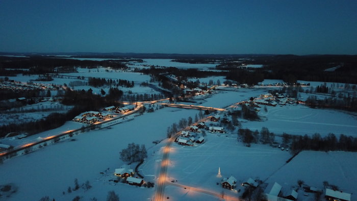 Kvällsfotografi av ett snötäckt landskap med belysta vägar och byggnader, taget från drönare.