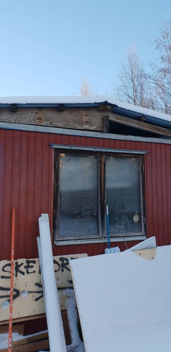 Gammal arbetsbod med slitna fönster och snö på taket, byggmaterial synligt i förgrunden.