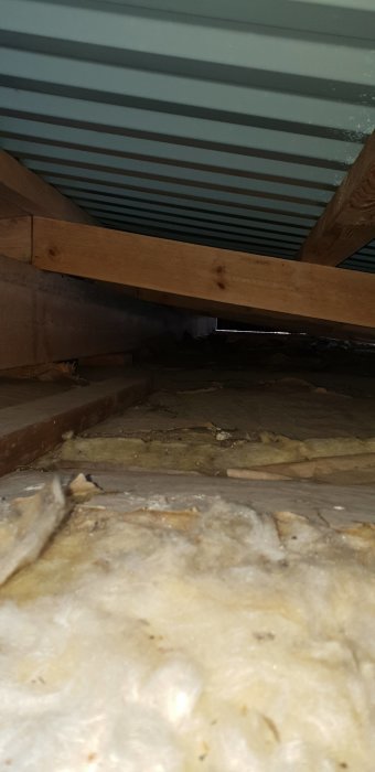 Vy under ett gammalt slitet innertak med exponerade takstolar och isoleringsmaterial, synligt skadat av fukt och ålder.